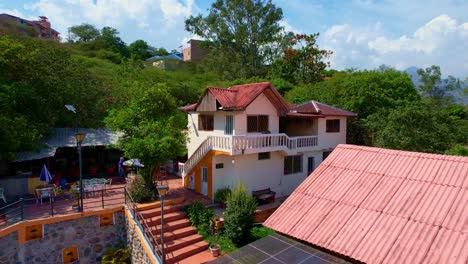 sunny day with blue sky over big green trees seen from a drone over flying a red roof farmhouse with blue pool with people enjoying the pool and the warmth of the sun