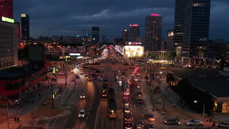 Forwards-fly-above-busy-multilane-road-leading-among-tall-office-buildings-in-city-centre.-Evening-shot-of-traffic-around-large-roundabout.-Warsaw,-Poland