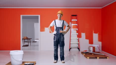 full body of asian man painter wearing safety helmet standing with arms akimbo looking at camera and smiling with painting wall in room