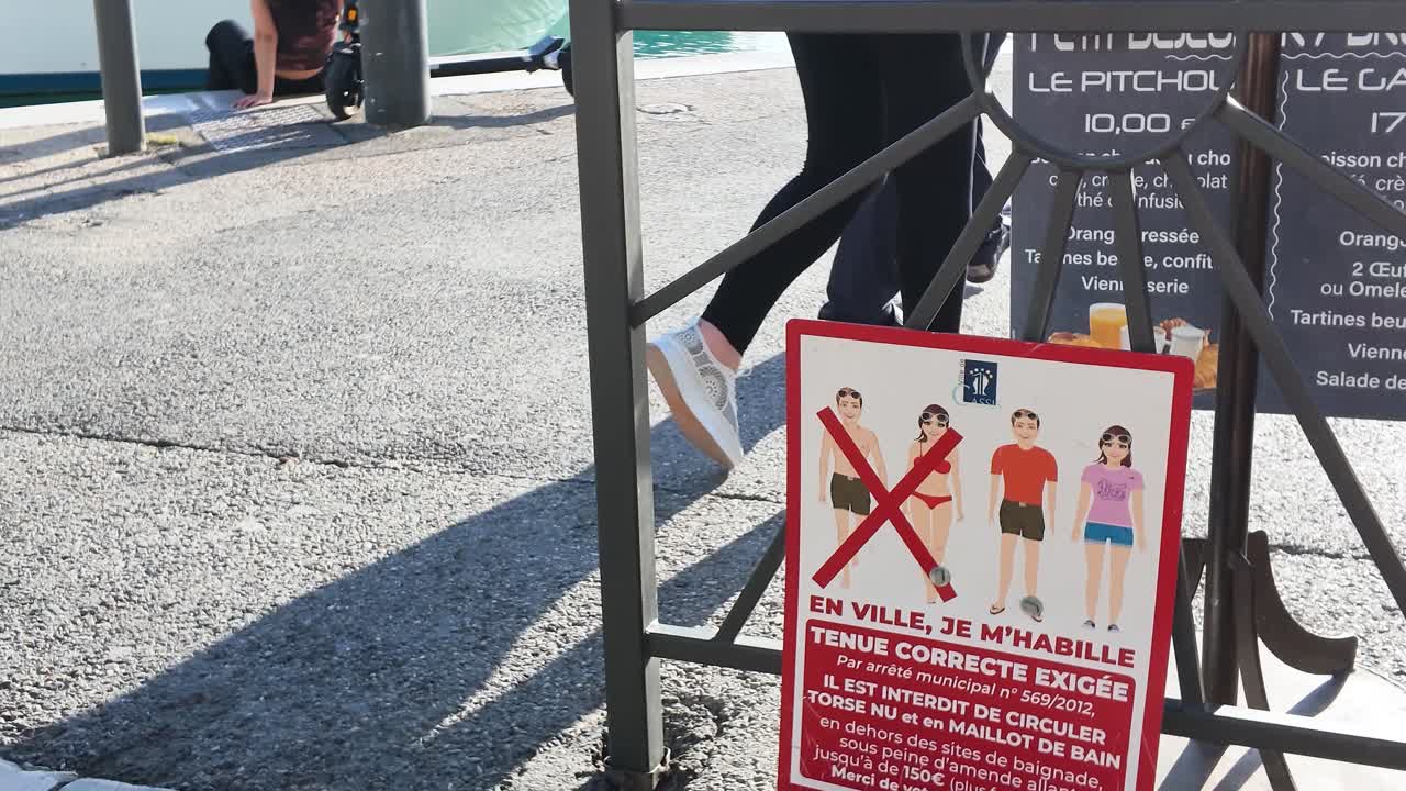 Un'inquadratura fissa mostra un cartello di regolamentazione francese che vieta i costumi da bagno su una strada soleggiata