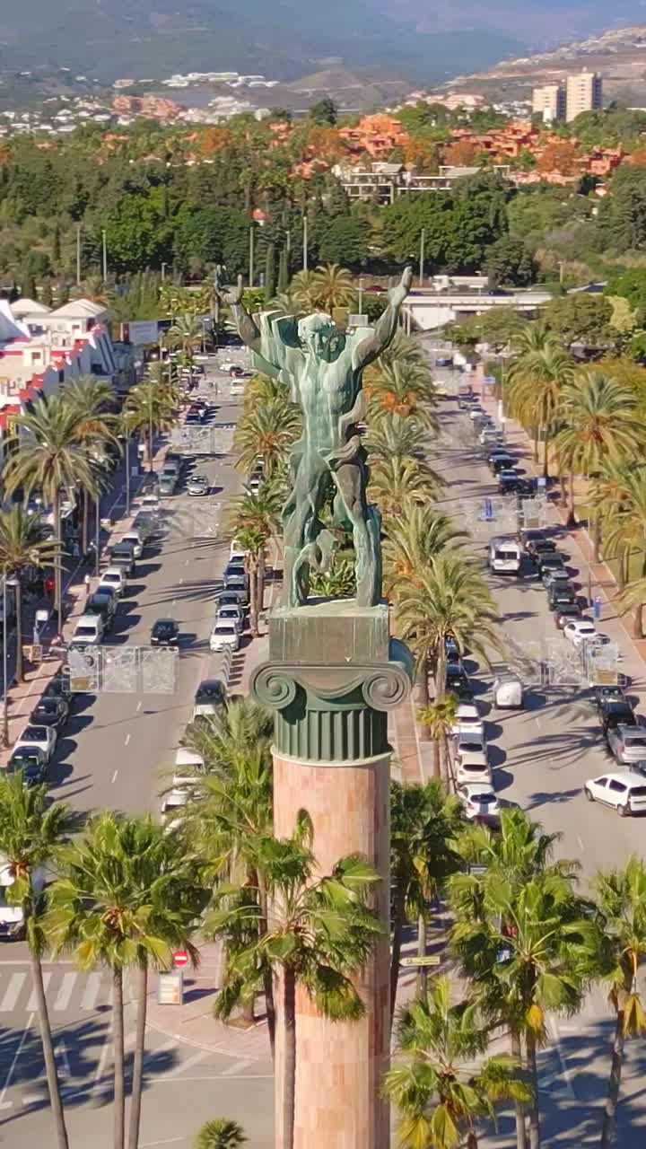 Orbita aerea verticale con allontanamento intorno alla statua de La Victoria a Puerto Banús, Marbella, in una giornata di sole splendente sulla Costa del Sol, Spagna.