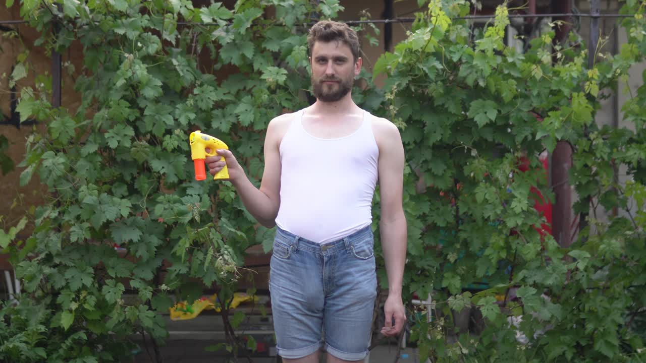 수염을 가진 유아적 인 젊은 남자가 서서 장난감 거품에서 으로 비누 거품을 발사합니다.