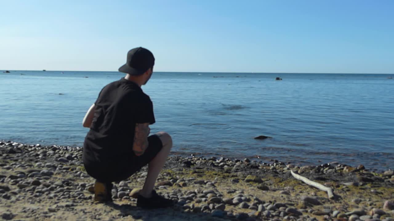 Slow motion video of a man in black clothing and tattoos skipping and throwing a flat rock on a sunny beach rocky and sandy shoreline during summer time. The rock bounces and skips on the blue ocean