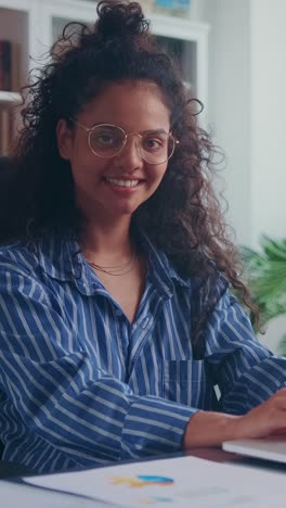 young indian woman looking at camera after using laptop sits in home office