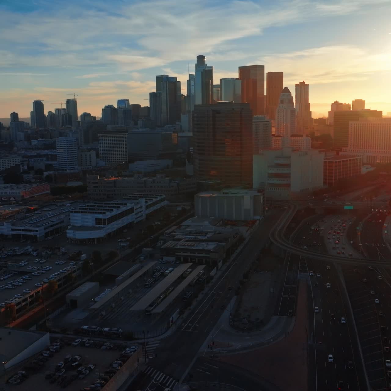 Multiple cars move by the roads and stand on the parking lots. Beautiful downtown skyscrapers of Los Angeles in the rays of setting sun