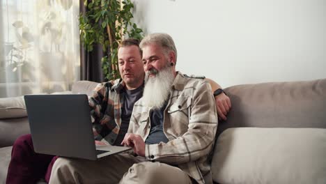 A-happy-middle-aged-man-with-gray-hair-and-a-lush-beard-sits-on-the-sofa-with-his-brunette-boyfriend-and-looks-at-the-monitor-of-a-gray-laptop-in-a-modern-apartment