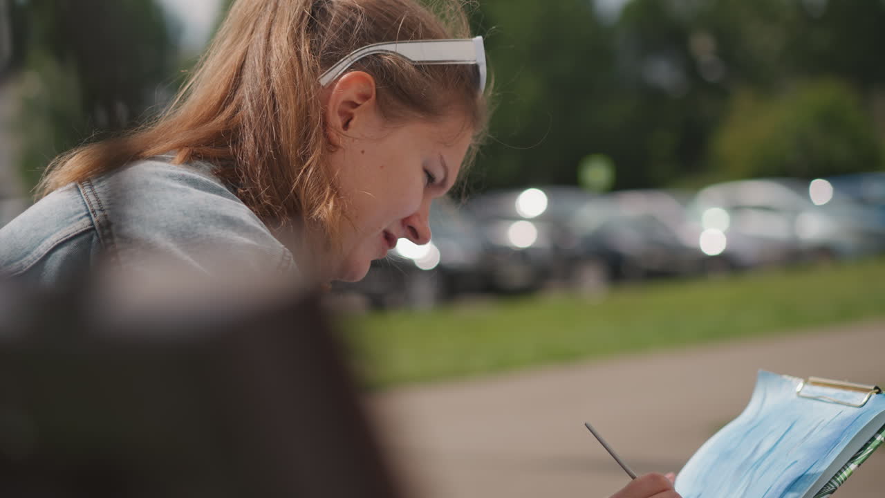 Młoda kobieta malująca na ławce w pobliżu parkingu, w dżinsowej kurtce i z kucykiem, zbliżenie profilu z pędzlem i papierem, samochody w tle z efektem bokeh, światło słoneczne podkreślające włosy, skupiona hobbystka uchwytująca kolor.