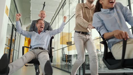 joyful business team celebrating success. four crazy happy colleagues riding chairs in office corridor, laughing, shouting and having fun