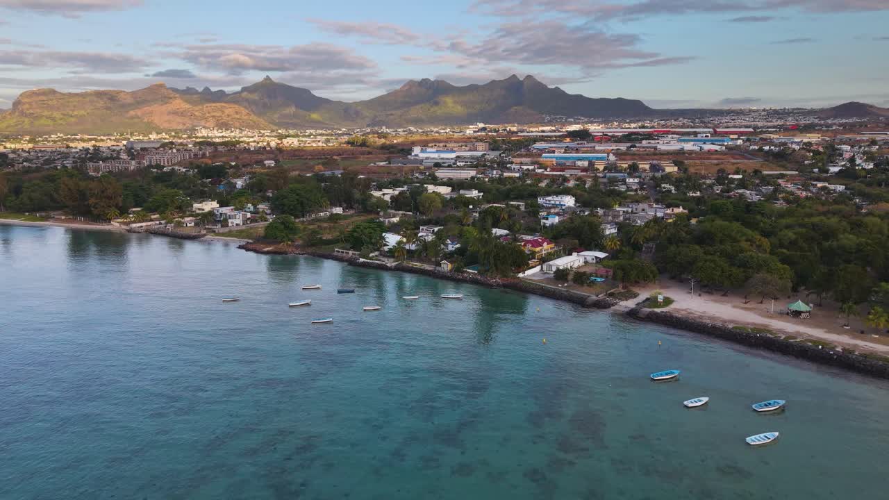 Pull in drone shot of a coastal resort near Port Louis, Mauritius, at sunset. Scenic tropical shoreline with palm trees, villas, and calm turquoise water captured in UHD