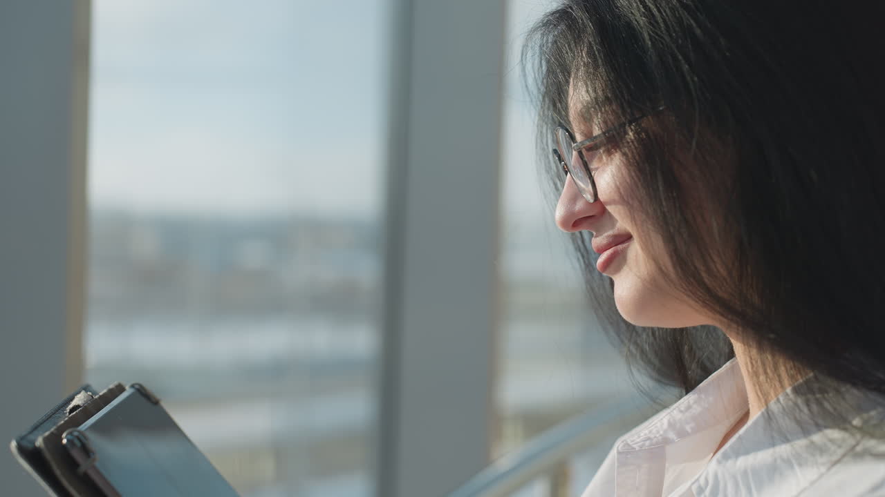 Close up side view of elegant woman with long dark hair and glasses smiling gently while chatting on tablet in bright indoor space with blurred window background and soft daylight