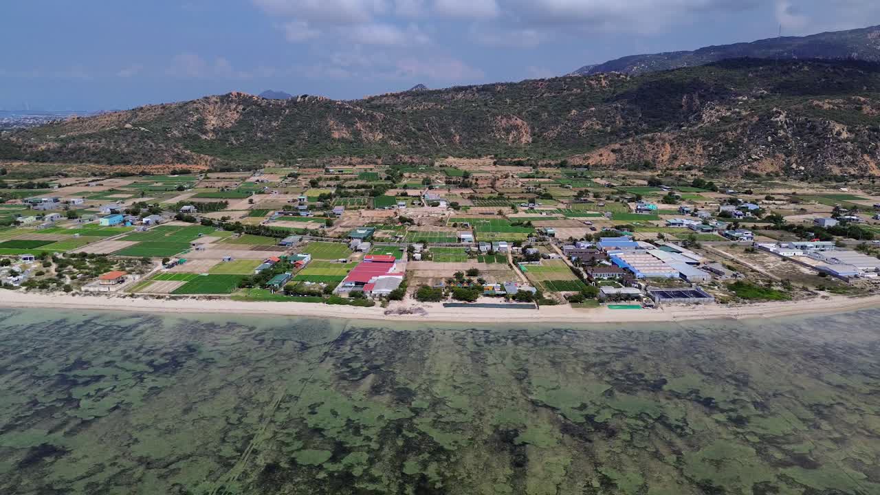 Drone footage flying from the sea towards the beach in Mỹ Hòa, Phan Rang. Right-to-left coastal motion reveals houses, shoreline, and soft waves on a sunny day.