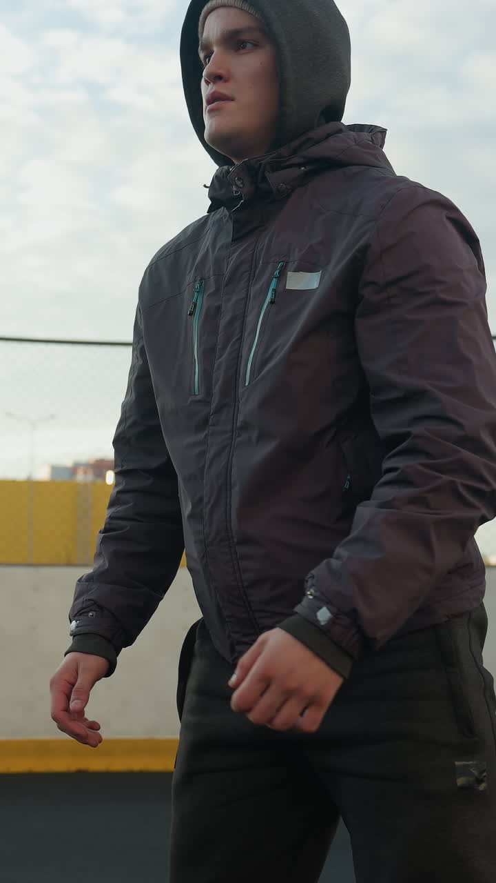 Close-up of focused sportsman performing deep squats with hands clasped during workout session on outdoor sports ground, yellow industrial building and wire fence in background