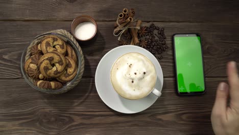 una mano femenina presiona la pantalla verde del teléfono inteligente tendido en la mesa con una taza de café.