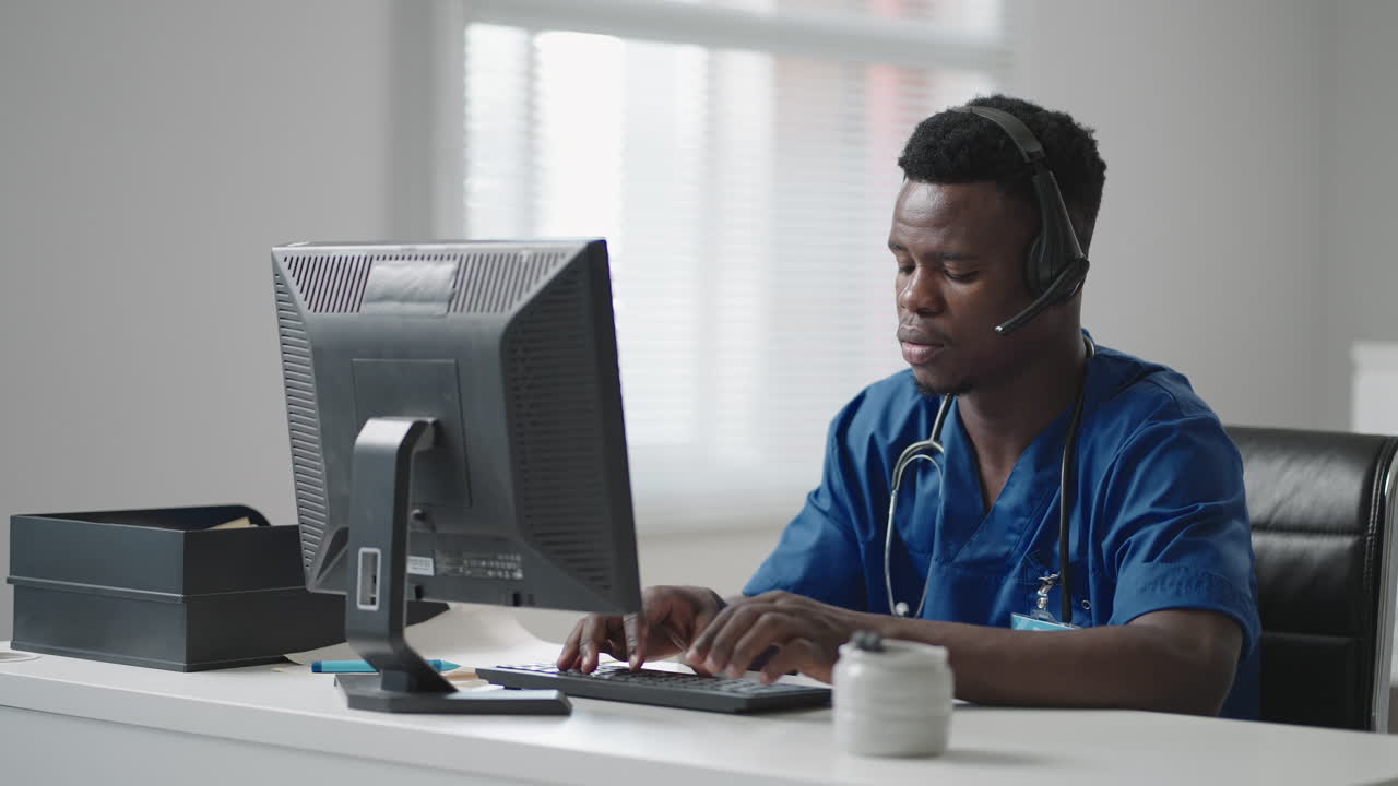 Un uomo afroamericano siede al computer in uniforme da medico e compila la cartella di un paziente mentre risponde a chiamate con le cuffie. La hotline delle ambulanze riceve chiamate e distribuisce ambulanze.