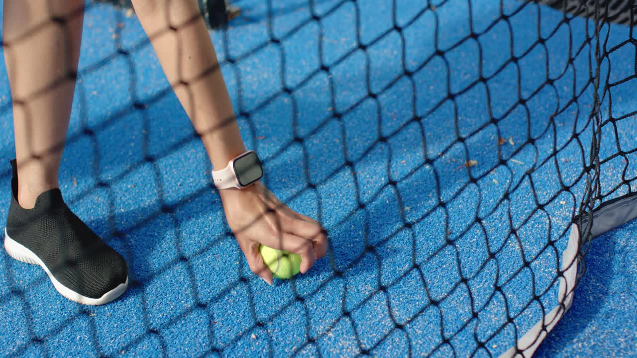 African American woman seeing tennis ball on blue court, reaching through net, picking up ball
