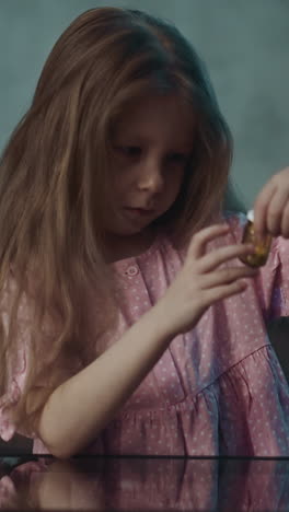 cute curious naughty little girl takes drug bottle full of white vitamin pills sitting at small glass table in semi-dark room at home close view slow motion