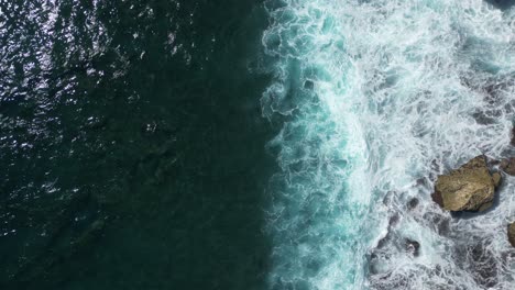 Birds-eye-view-of-waves-hitting-rocks-in-the-South-of-Bali,-Indonesia-on-a-sunny-afternoon