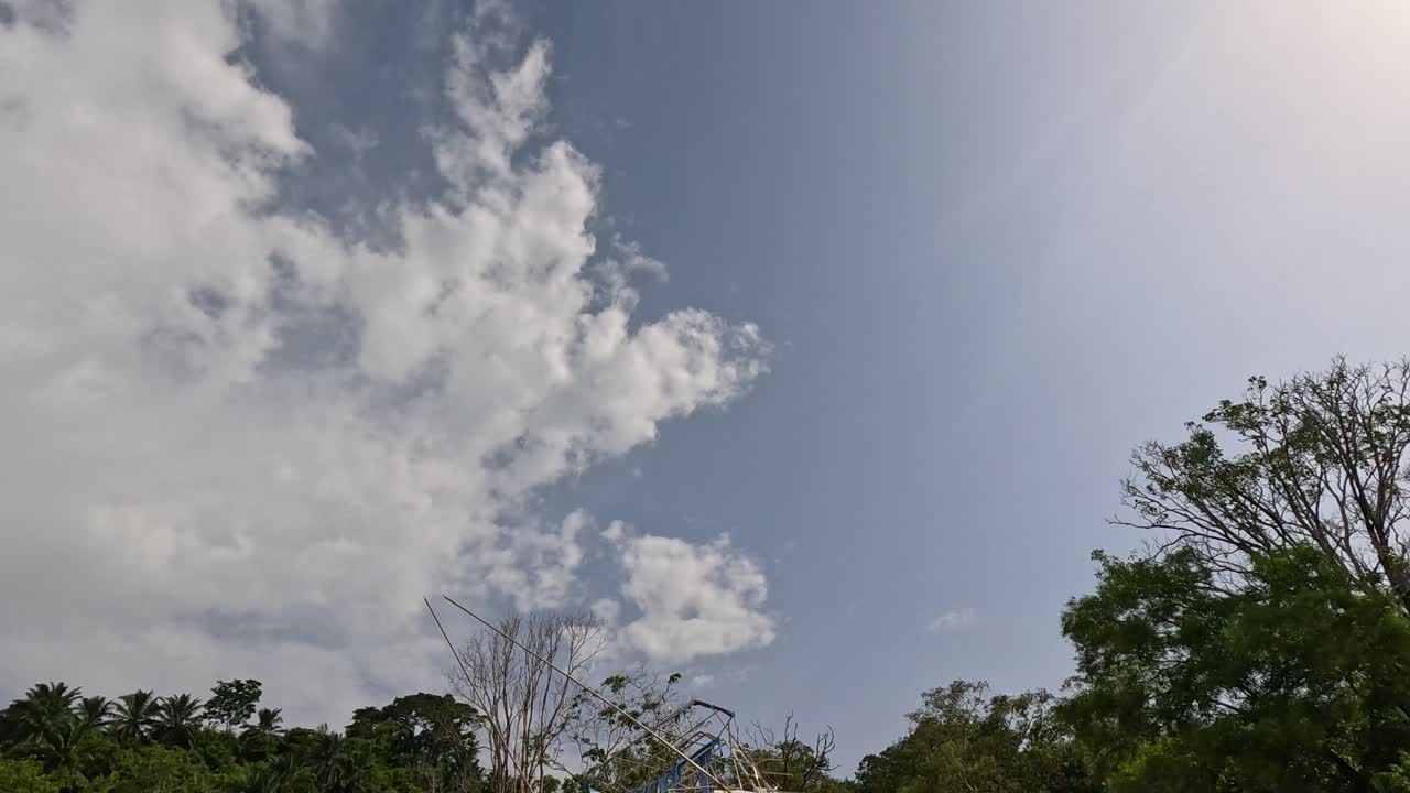A stationary, beached boat rests on a quiet sandy shore with lush green trees and partly cloudy skies, captured in a slow panning shot with natural daylight