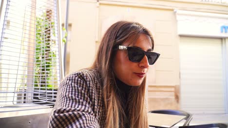 chica caucásica con cabello lacio, gafas de sol, labios rojos y un abrigo a cuadros, sentada en una terraza junto a una estufa y bebiendo de un vaso