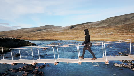 beautiful tracking shot of a person, passing a narrow wooden bridge leading over a river flowing through a stony, barren landscape, sunny day, outdoor, wilderness