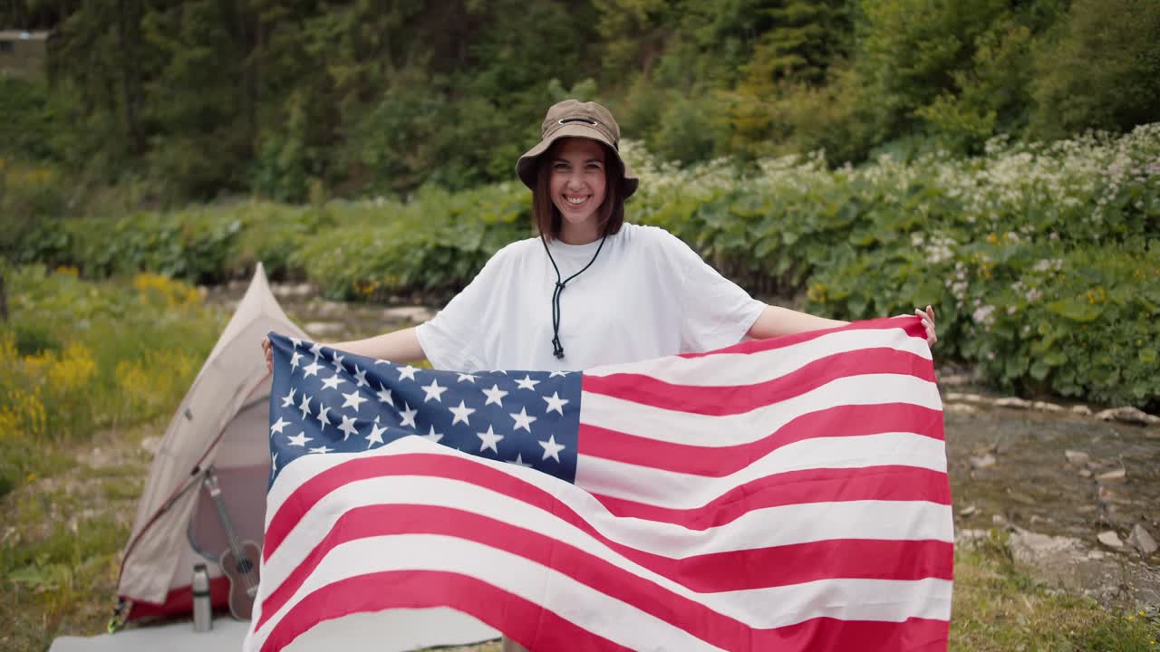 retrato de una chica morena patriótica con una camiseta blanca que se para con la bandera de los estados unidos de américa sonriendo contra el telón de fondo de un bosque verde y un río de montaña