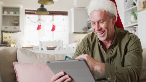 Glücklicher-Kaukasischer-Mann-Mit-Weihnachtsmannmütze-Und-Tablet-Für-Videoanrufe-Im-Wohnzimmer