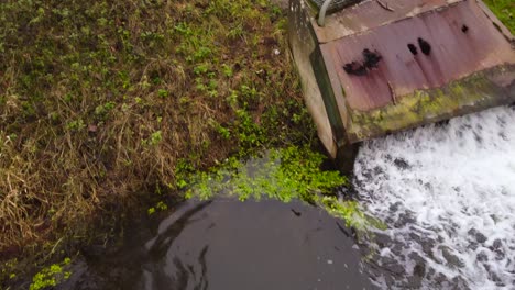 Water-pollution,-factory-sewage-water-flowing-out-on-river-from-rusty-pipeline---dolly-backward-tilt-down-shot