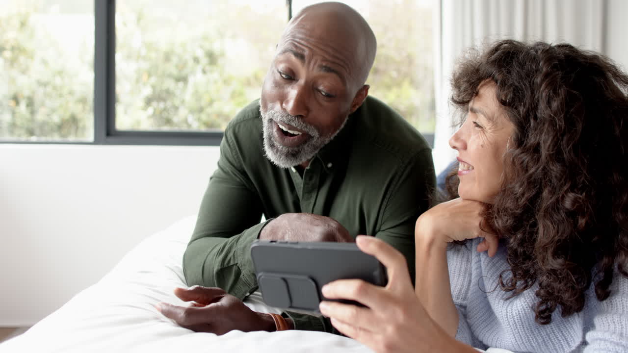 una pareja feliz, madura y diversa acostada en la cama usando un teléfono inteligente en casa, en cámara lenta
