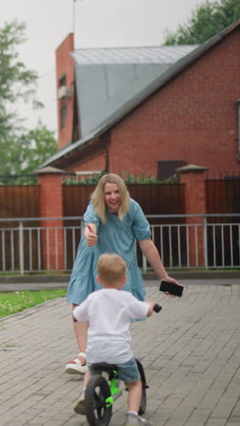a woman happily reaches out to her younger child riding a bicycle, offering support and encouragement, the older sibling rides from behind, with a building behind them
