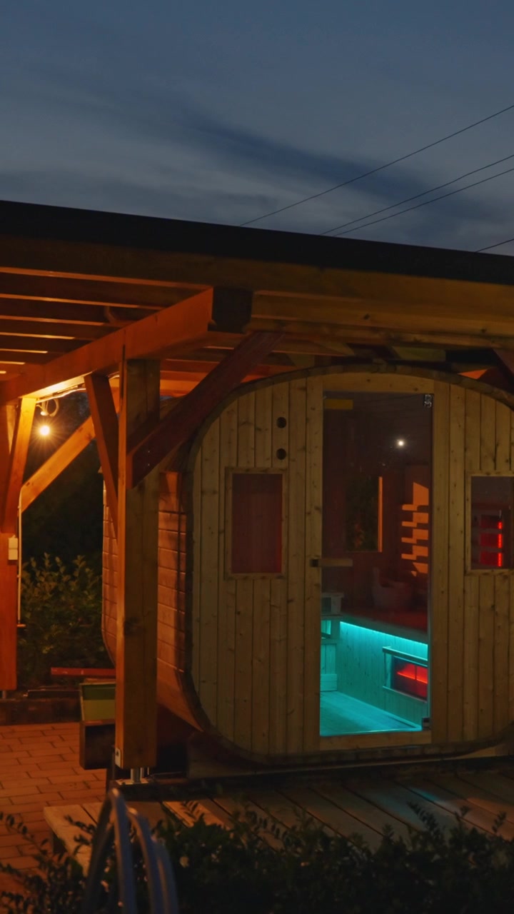 L'illuminazione calda mette in risalto una moderna sauna a botte in legno al tramonto. Atmosfera invitante e tranquilla, perfetta per il relax. Video verticale accogliente ideale.