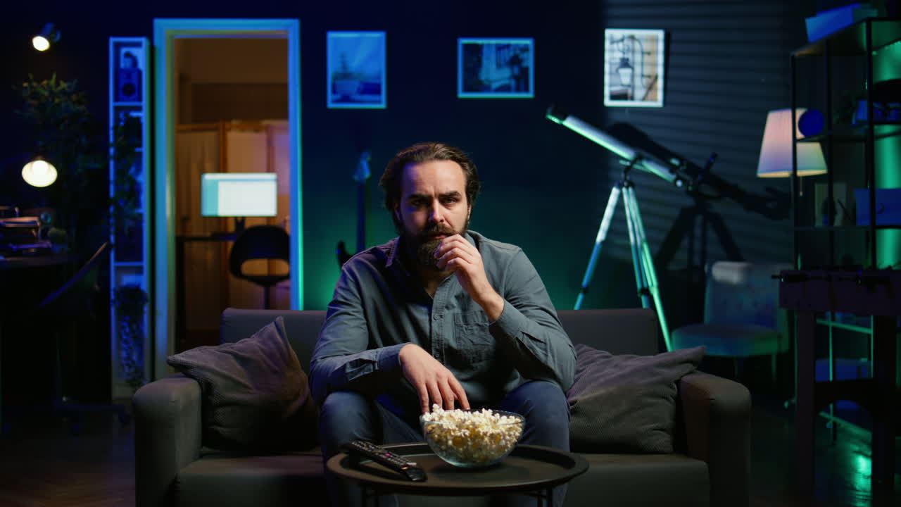 hombre desconcertado comiendo palomitas de maíz mientras ve una película confusa en la televisión