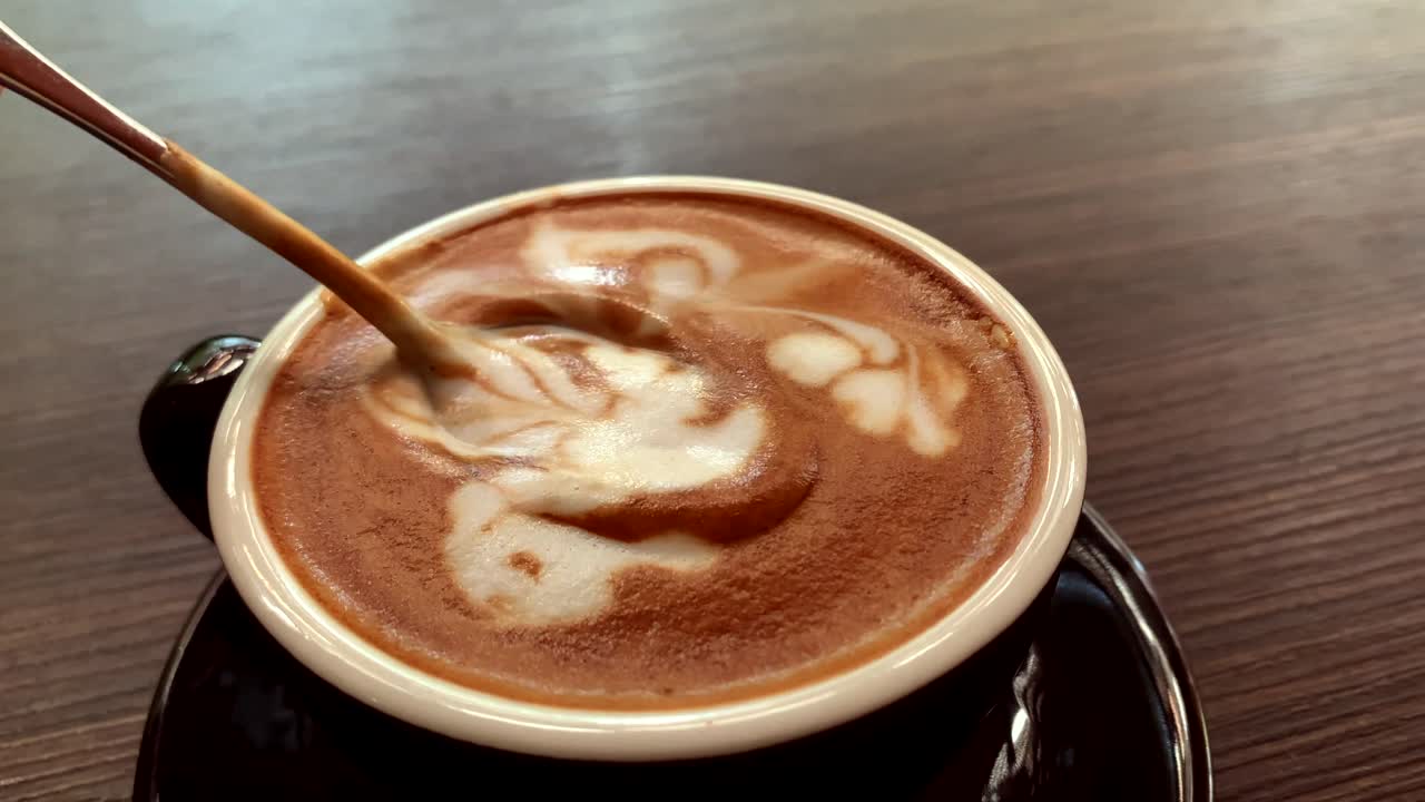 A person's hand, no face is shown, steering the foamy cafe latte from a cup of coffee that is placed on the table