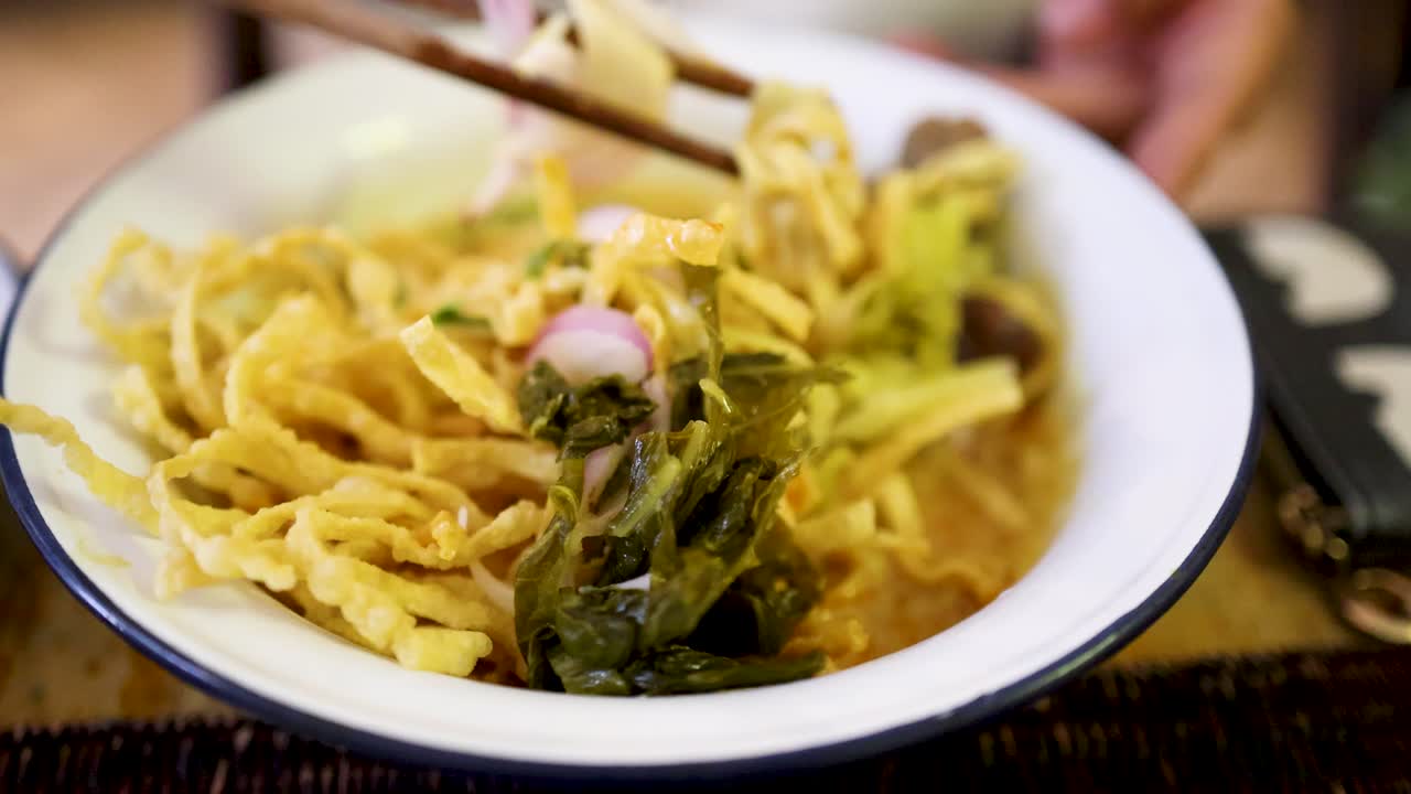 Hand uses chopsticks to lift crispy noodles and pickled greens from a bowl of Northern Thai beef curry noodles, under warm indoor lighting