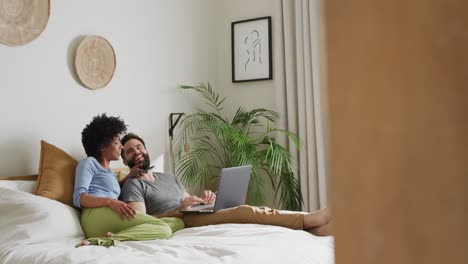 Video-De-Una-Feliz-Pareja-Diversa-Relajándose-En-Casa,-Acostada-En-La-Cama-Usando-Una-Computadora-Portátil-Y-Un-Teléfono-Inteligente,-Copiando-Espacio