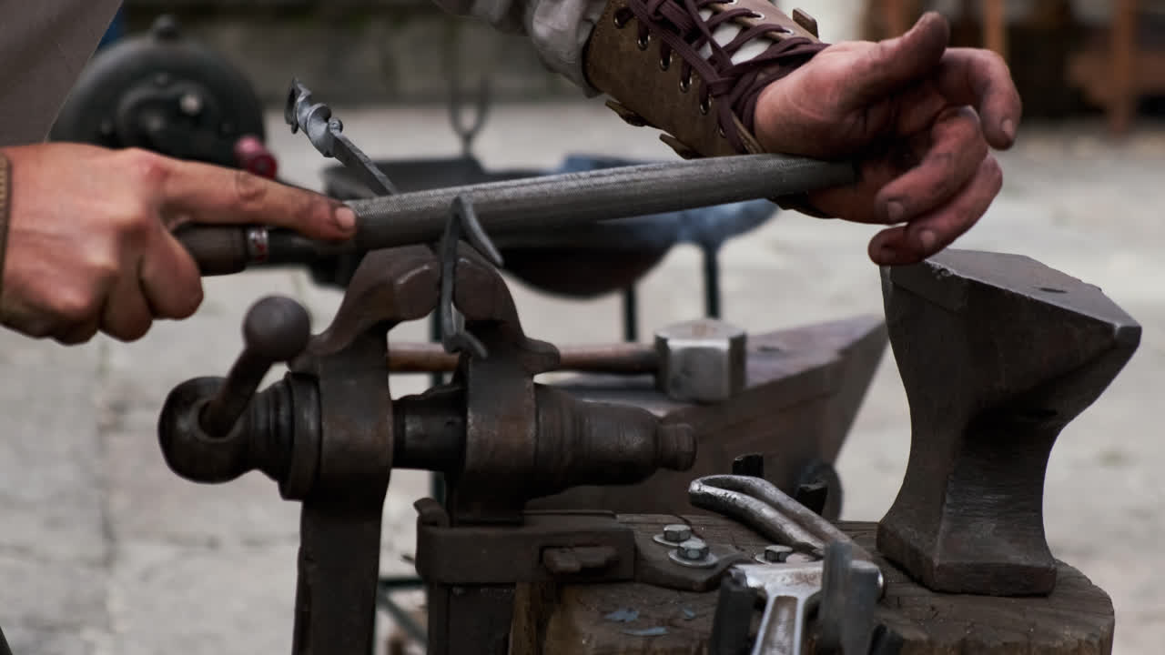 Static lateral close-up of a blacksmith smoothing a clamped metal piece with a hand file; anvil and hammer in soft background, assorted tools around the workbench