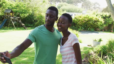 smiling african american couple taking selfie with smartphone embracing in sunny garden