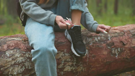 close-up of woman with long nails tying her shoelace while sitting on fallen tree during hike, she wears a gray sweater and light blue jeans, focused on adjusting her black sneakers in forest setting