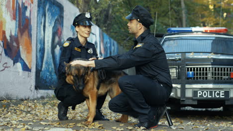 Cheerful-Couple-Of-Policeman-And-Policewoman-Sitting-At-The-Street-And-Petting-Shepherd-Dog