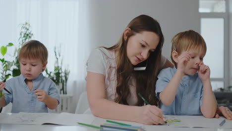 A-young-mother-with-two-children-talking-on-a-mobile-phone-draws-with-a-pencil-and-helps-children-draw-with-colored-pencils