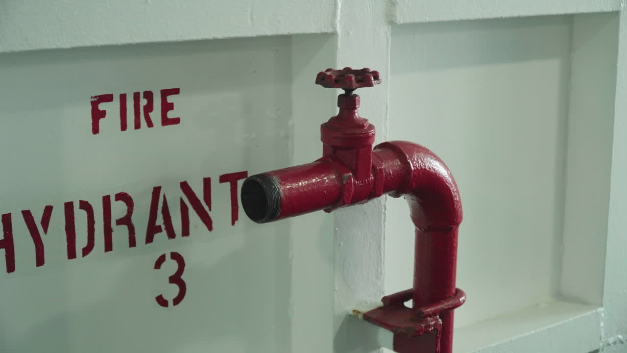 Idrante antincendio rosso montato a parete, primo piano ravvicinato di valvola e tubo lucidi su un pannello bianco verniciato con la scritta a stencil "IDRANTE ANTINCENDIO". L'immagine evoca un'atmosfera di sicurezza industriale, ispezione e manutenzione, con texture metallica, bulloni e leggera ruggine.