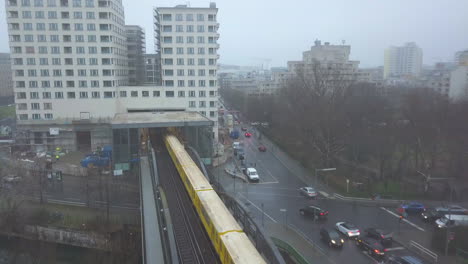 Aerial-follow-Shot-of-Yellow-Subway-Train-on-Bridge-in-Berlin-entering-Train-Station,-Foggy-weather-with-clouds,-Drone-Shot