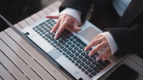 close-up of hands with delicate rings and polished nails typing on laptop keyboard outdoors, placed on wooden table with smartphone nearby, creating an aesthetic scene of modern work and style