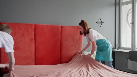 Eine-Brünette-Putzfrau-In-Einem-Weißen-T-Shirt-Und-Einer-Blauen-Schürze-Macht-Zusammen-Mit-Ihrem-Blonden-Männlichen-Kollegen-Das-Bett-Mit-Einer-Rosa-Decke-Und-Legt-Graue-Kissen-Auf,-Während-Sie-Das-Schlafzimmer-Putzt