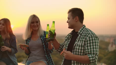 Two-couples-in-love-clink-glasses-of-beer-on-the-roof-at-sunset-and-eat-pizza-at-a-friend's-party