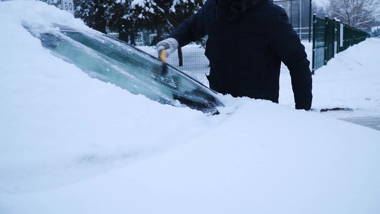 Den unge mannen rengör bilens framruta från snön med en gul borste på vintern.