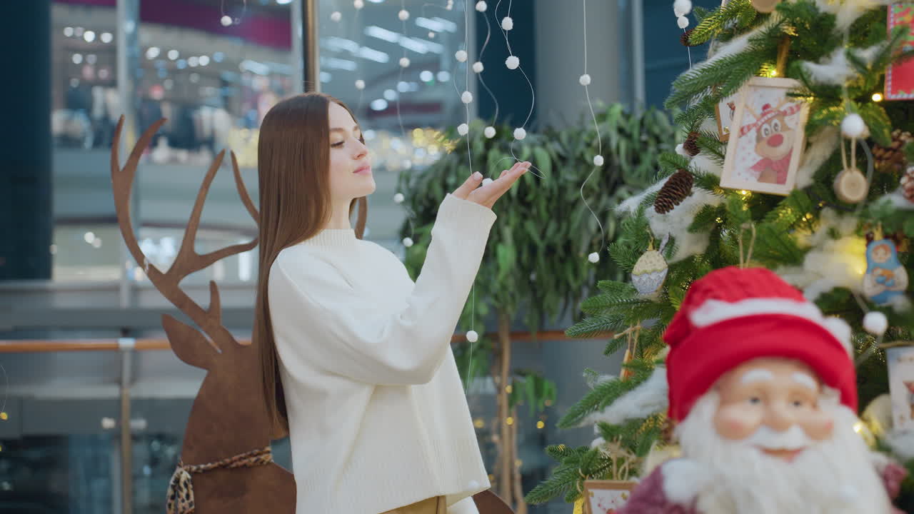 Woman in white sweater admires Christmas tree decorations in well-decorated mall, evoking festive spirit with ambient lights and ornaments
