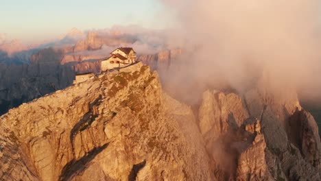 Luftdrohnenaufnahme-Einer-Roten-Bergwand-Mit-Fliegenden-Wolken-Bei-Sonnenuntergang-Und-Hütte-Rifugio-Nuvolau-Auf-Dem-Gipfel