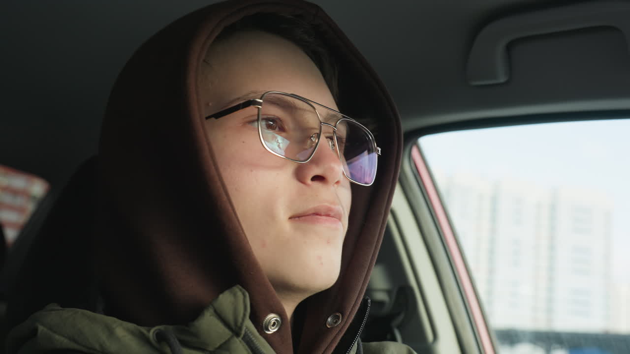 close up portrait of hoodie clad youth inside parked car peering through glass at clear blue sky while sun light reflects off glasses with crisp winter urban view in background