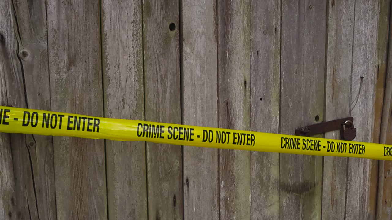 Yellow colored police line foresnic investigation tape ribbon pulled in front of a farm house or barn shed wooden door with a rusty padlock locking the door during day time. bokeh blurry background