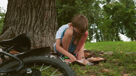 a young child squats under a large tree with a bicycle lying close by, he is deep in thought, with his hand resting on his leg